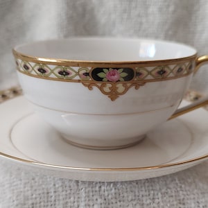May include: A white teacup and saucer with a gold rim and floral design. The cup has a black and gold band with a pink rose and green leaves. The saucer has a similar design.