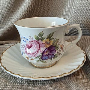 May include: A white teacup and saucer set with a floral design featuring pink, purple, and yellow flowers. The cup and saucer have gold trim. The saucer has a scalloped edge.