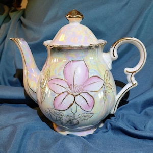 May include: A white porcelain teapot with a pink floral design and gold accents. The teapot has a pearlescent glaze and a delicate floral pattern.