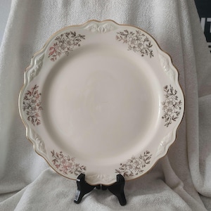 May include: A white ceramic dinner plate with a gold floral design around the rim. The plate has a scalloped edge and is on a black stand.