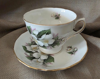 May include: A white teacup and saucer set with a floral design. The teacup and saucer are decorated with white flowers, green leaves, and pink accents. The edges of the teacup and saucer are trimmed with gold.