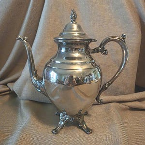 May include: A silver-colored tea or coffee pot with a lid and a curved handle. The pot has a bulbous body and sits on three ornate feet. The surface reflects the surrounding environment. The spout is curved and elegant.