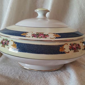 May include: A covered serving dish with a white base and lid, featuring a blue and cream striped design. The dish is adorned with floral basket motifs and gold trim. It has two handles and a knob on the lid.