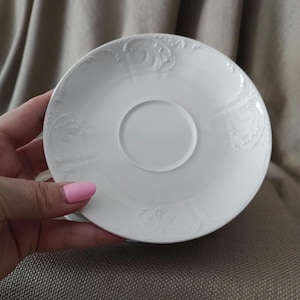 May include: White porcelain saucer with a raised, ornate design around the edge.