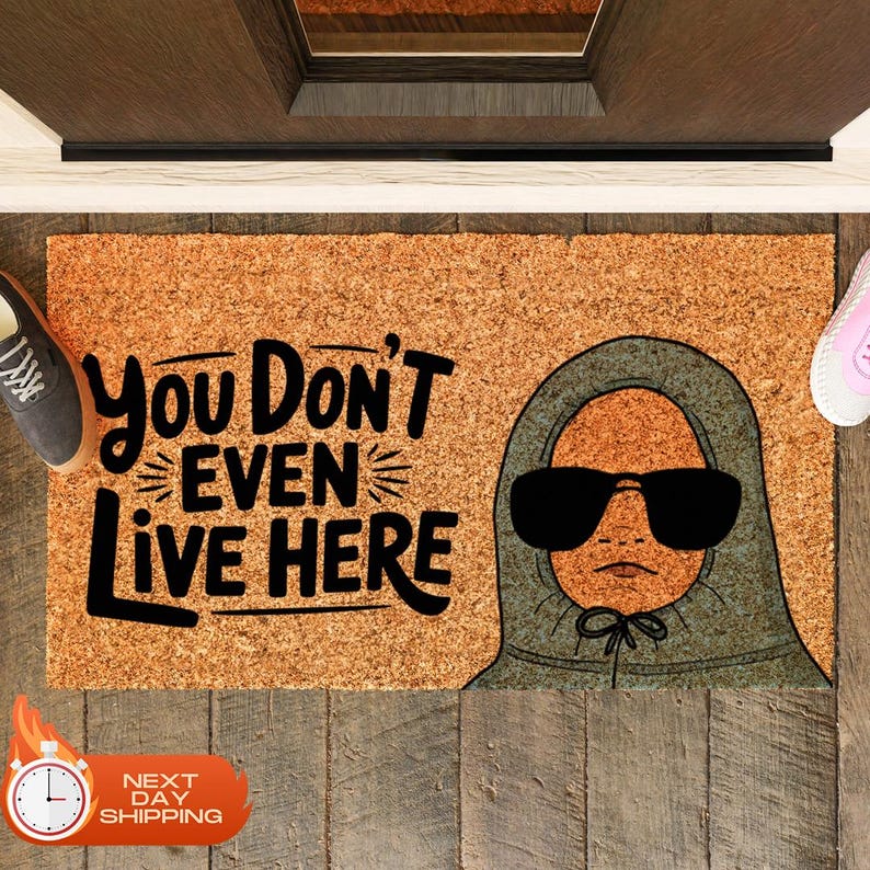 Puede incluir: Una alfombra de bienvenida marr&oacute;n con el texto "You Don't Even Live Here" en letras negras. Una ilustraci&oacute;n de dibujos animados de una persona con una sudadera con capucha gris y gafas de sol est&aacute; en el lado derecho de la alfombra.