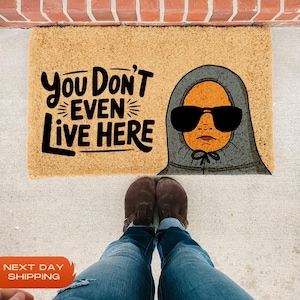 Puede incluir: Una alfombra de bienvenida marr&oacute;n con el texto "You Don't Even Live Here" en letras negras. Una ilustraci&oacute;n de dibujos animados de una persona con una sudadera con capucha gris y gafas de sol est&aacute; en el lado derecho de la alfombra.