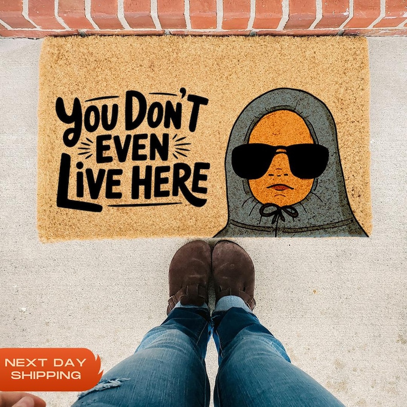 Puede incluir: Una alfombra de bienvenida marr&oacute;n con el texto "You Don't Even Live Here" en letras negras. Una ilustraci&oacute;n de dibujos animados de una persona con una sudadera con capucha gris y gafas de sol est&aacute; en el lado derecho de la alfombra.