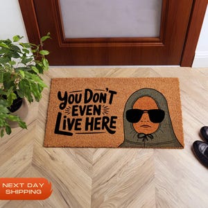 Puede incluir: Una alfombra de coco marr&oacute;n con el texto "You Don't Even Live Here" en letras negras. Una ilustraci&oacute;n de dibujos animados de una persona con una sudadera con capucha verde y gafas de sol est&aacute; en el lado derecho de la alfombra.