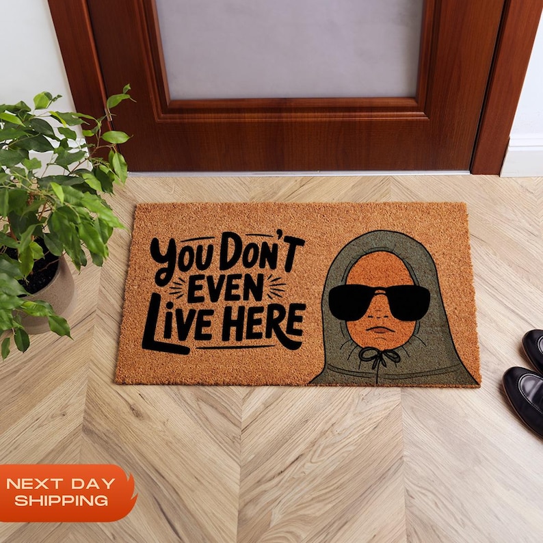 Puede incluir: Una alfombra de coco marr&oacute;n con el texto "You Don't Even Live Here" en letras negras. Una ilustraci&oacute;n de dibujos animados de una persona con una sudadera con capucha verde y gafas de sol est&aacute; en el lado derecho de la alfombra.