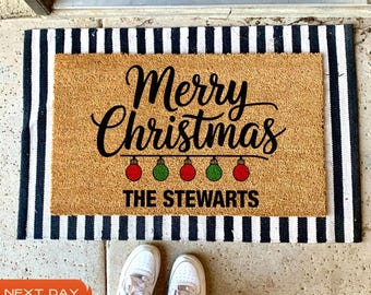 May include: A brown coir doormat with black and white stripes around the edges. The doormat has the text "Merry Christmas" in black cursive font with a string of five red and green Christmas ornaments hanging below. The text "The Stewarts" is printed below the ornaments.