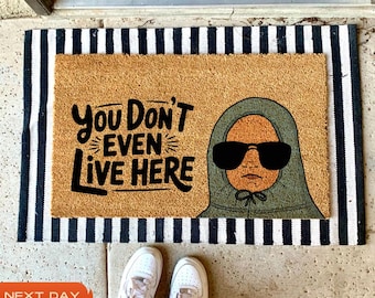 May include: A black and white striped doormat with the text "You Don't Even Live Here" in black lettering. A cartoon illustration of a person wearing a green hooded sweatshirt and sunglasses is on the doormat.