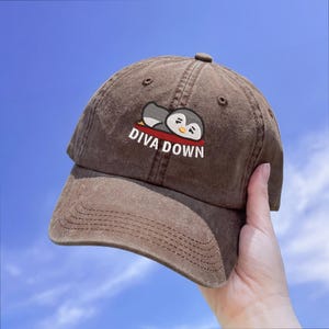 May include: Brown baseball cap with a curved brim. The cap features an embroidered design of a cartoon penguin and the text "DIVA DOWN" in white. The cap is made of a soft, textured fabric.