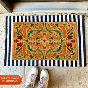 May include: A rectangular doormat with a central design of orange, green, and brown floral patterns. The mat is bordered by a black and white striped pattern. White sneakers are visible at the bottom of the image.