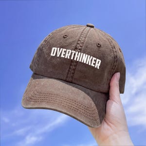 Könnte beinhalten: Braune Baseballkappe mit dem Wort "OVERTHINKER" in weißen Blockbuchstaben. Die Kappe wird vor blauem Himmel gehalten. Die Kappe hat einen gebogenen Schirm und einen Knopf oben.