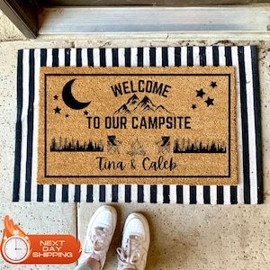 May include: A brown coir doormat with the text "Welcome to our campsite" and the names "Tina & Caleb". The doormat features a black silhouette of mountains, a crescent moon, stars, and camping chairs around a campfire.