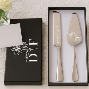 May include: A silver cake knife and server set with floral and initial details, presented in a black box. The knife is engraved with a name and the server with initials. A blank card and a vase of white roses are also visible.