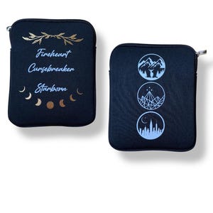 May include: Two black zippered pouches with different designs. One pouch has gold lettering that reads "Fireheart Cursebreaker Starborn" with a gold floral design above the text. The other pouch has three white circles with different designs. The top circle has a mountain range with a deer in the centre. The middle circle has a crystal with three stars above it. The bottom circle has a city skyline.