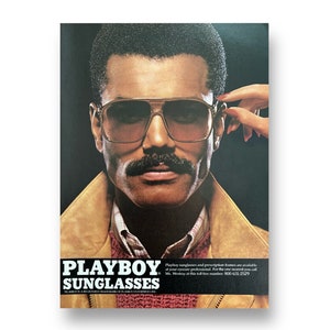 A vintage Playboy Sunglasses ad shows a man in sunglasses and a mustache. A hand with red nails reaches over to take the glasses off. The man is wearing a red sweater and suede 80s jacket. Beneath him reads, Playboy Sunglasses.