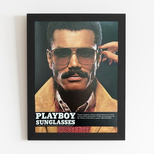 A vintage Playboy Sunglasses ad is in a black from. The ads shows a man in sunglasses and a mustache. A hand with red nails reaches over to take the glasses off.