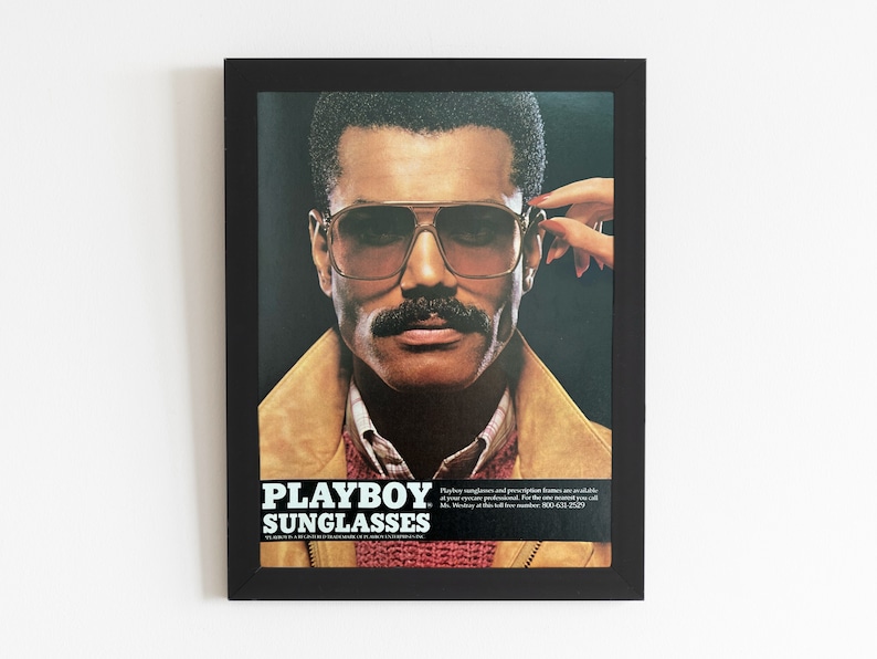 A vintage Playboy Sunglasses ad is in a black from. The ads shows a man in sunglasses and a mustache. A hand with red nails reaches over to take the glasses off.