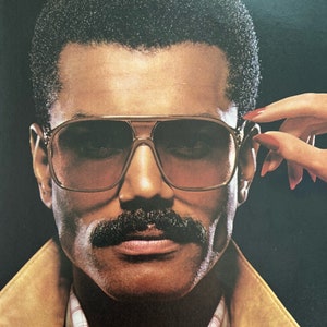 A vintage Playboy Sunglasses ad shows a man in sunglasses and a mustache. A hand with red nails reaches over to take the glasses off. The man is wearing a red sweater and suede 80s jacket. Beneath him reads, Playboy Sunglasses.