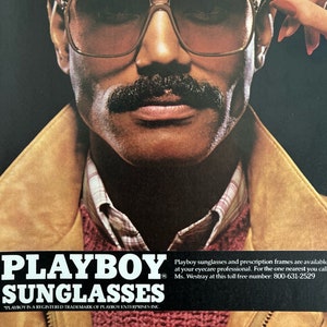 A vintage Playboy Sunglasses ad shows a man in sunglasses and a mustache. A hand with red nails reaches over to take the glasses off. The man is wearing a red sweater and suede 80s jacket. Beneath him reads, Playboy Sunglasses.