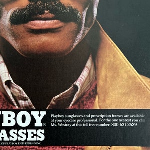 A vintage Playboy Sunglasses ad shows a man in sunglasses and a mustache. A hand with red nails reaches over to take the glasses off. The man is wearing a red sweater and suede 80s jacket. Beneath him reads, Playboy Sunglasses.