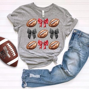 May include: A gray t-shirt with a pattern of footballs and bows. The bows are red and black. The footballs are brown and white. The shirt is on a white surface with a pair of blue jeans.