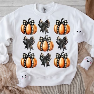 May include: White sweatshirt featuring a repeating pattern of orange pumpkins with black and white checkered bows. The design includes solid black bows. Small white ghost plushies are placed around the sweatshirt.