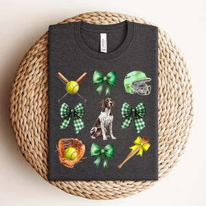 May include: Dark gray t-shirt featuring a watercolor design with baseball and softball elements. Includes a German Shorthaired Pointer, green bows, a softball helmet, and a baseball glove. The shirt has a crew neck.