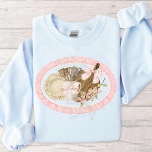 Op de afbeelding: Lichtblauw sweatshirt met een aquarelontwerp van keukenartikelen. Het ontwerp omvat een bord, keukengerei, een snijplank met een roze strik en bessen, alles binnen een roze ovale lijst. De sweatshirt heeft lange mouwen.