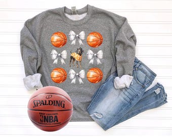 May include: A gray sweatshirt with a basketball and bow design. The sweatshirt has a basketball, bow, and dog graphic. The sweatshirt is paired with blue jeans.