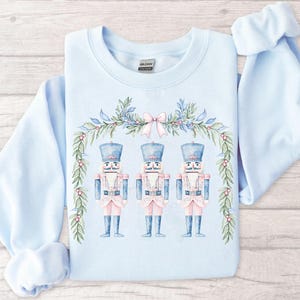 May include: A light blue sweatshirt with a watercolor design of three nutcracker soldiers standing under a pink and green garland. The nutcrackers are wearing blue and pink uniforms with gold accents.