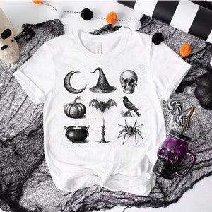 May include: White t-shirt with a black and white graphic print of Halloween icons, including a crescent moon, witch's hat, skull, bat, crow, pumpkin, cauldron, candle, and spider.
