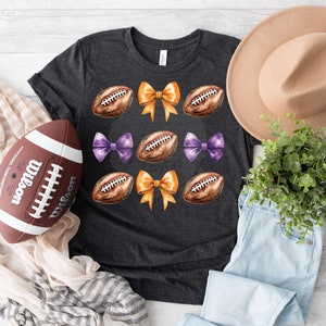 May include: A dark grey t-shirt featuring a football and bow design. The footballs are brown and white, and the bows are purple and orange.