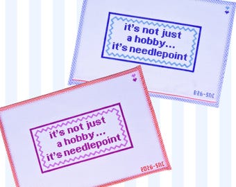 Op de afbeelding: Twee rechthoekige naaldpuntcanvassen met de tekst "it's not just a hobby... it's needlepoint" in een zigzagrand. Eén canvas heeft blauwe tekst en een blauw geblokte rand, de andere paarse tekst en een rood geblokte rand.