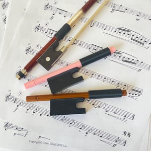 May include: Four violin bows and bow holders are arranged on sheet music. The bows are in shades of brown, pink, and natural wood, with black and gold details. The bow holders are black. The sheet music features musical notation and text.