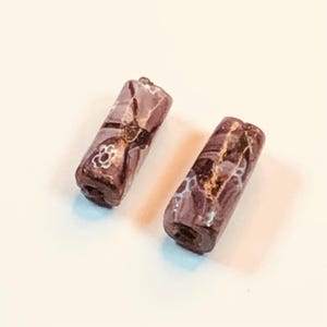 May include: Two cylindrical beads with a dark purple base color and intricate patterns in white and gold. One bead features a small white floral design. The beads are likely for jewelry making.