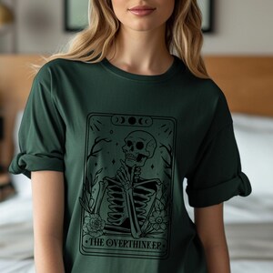 Może przedstawiać: Zielony T-shirt z czarno-białym rysunkiem szkieletu trzymającego kartę tarota. Karta zawiera napis "The Overthinker".