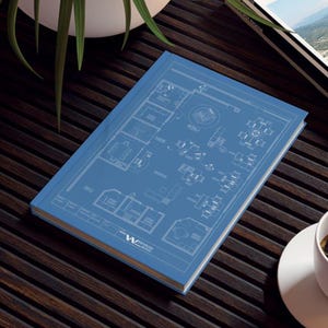 Könnte beinhalten: Ein blaues Notizbuch mit einem detaillierten architektonischen Bauplan-Design. Das Cover zeigt einen Grundriss mit weißen Linien und Text. Das Notizbuch liegt auf einer dunklen Holzoberfläche, mit einer weißen Tasse und Untertasse.