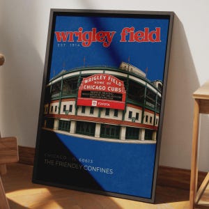 Puede incluir: Un póster enmarcado de Wrigley Field en Chicago, que muestra el exterior del estadio con el texto rojo Wrigley Field Est. 1914, un letrero rojo y la inscripción Chicago IL 60613 The Friendly Confines sobre un fondo azul intenso.