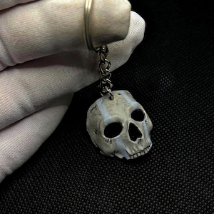 Può includere: Un portachiavi a forma di teschio con catena e anello argentati. Il teschio è grigio con accenti bianchi e presenta orbite oculari aperte e una mascella dettagliata. Lo sfondo è nero.