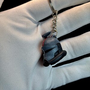May include: A gray and black keychain featuring a miniature helmet design. The helmet has a visor and other details. The keychain is attached to a silver metal ring and chain. The background is black.