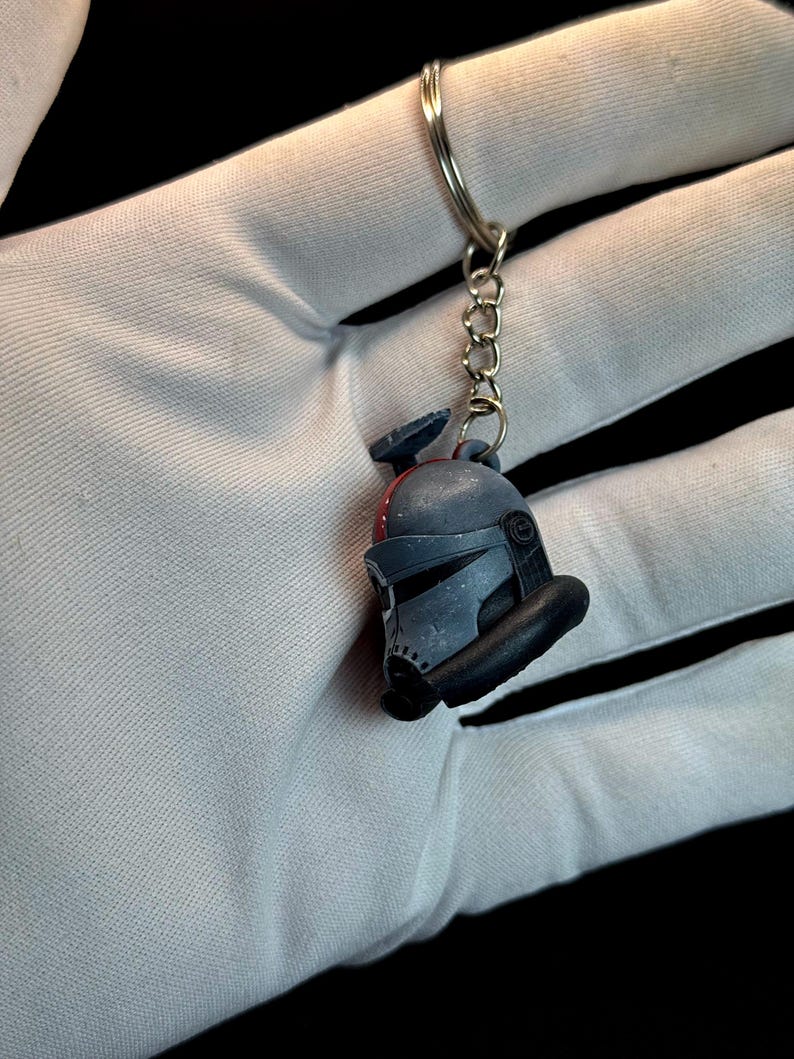 May include: A gray and black keychain featuring a miniature helmet design. The helmet has a visor and other details. The keychain is attached to a silver metal ring and chain. The background is black.
