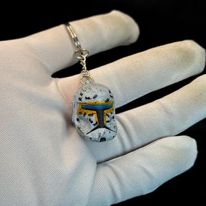 May include: A small, detailed keychain featuring a miniature helmet with a weathered, battle-worn appearance. The helmet is primarily white with black, yellow, and blue accents. The keychain is attached to a silver-colored chain.