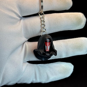 Puede incluir: Un llavero negro con la cabeza de un personaje con rostro rojo y capucha negra. El llavero está sujeto a una cadena y un anillo plateados. La cara del personaje tiene un diseño rojo.