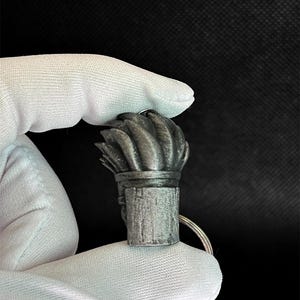 May include: A metallic gray keychain charm featuring a sculpted, textured top section with swirling, flame-like ridges and a cylindrical base with a vertical, wood-grain-like pattern. A small silver metal ring is attached to the side of the object.