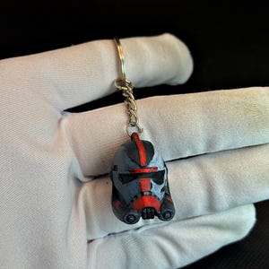 May include: A small, detailed keychain featuring a helmet design in shades of gray, black, and red. The helmet has a distinctive visor and markings, attached to a silver-colored chain and keyring. The keychain is held against a white fabric background.