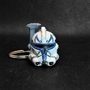May include: A white and blue miniature helmet keychain with a gold-colored keyring. The helmet has blue markings and is attached to a gray support. The background is black.