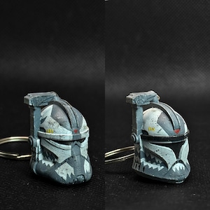 May include: A miniature, weathered, gray and white helmet keychain. The helmet features a distinctive design with angular shapes and a visor. The keychain is attached to a silver-colored ring. The overall aesthetic suggests a military or sci-fi theme.